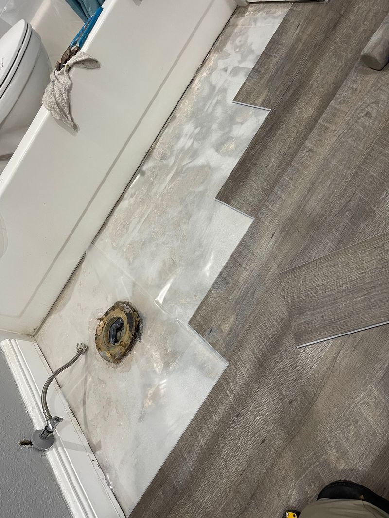 Gray vinyl plank being fitted around the toilet flange in a bathroom
